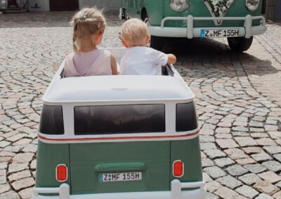 Zwei kleine Kinder im Kinderbulli auf einer Hochzeit im gepflasterten Hof Elektroauto Kinderprogramm VW Oldtimer